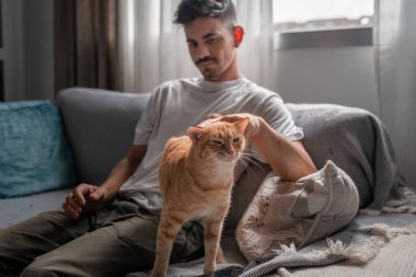 Kanepede uzanmış genç bir adam kahverengi bir tekir kediyle etkileşime geçiyor.