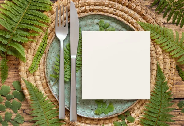 Square blank paper card on table setting decorated with fern leaves on wooden table top view. Tropical mock-up scene with invitation card flat lay