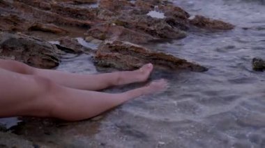 Genç bir kadının kayalık sahilde oturması, suya ayak vurması, okyanus dalgalarının tadını çıkarması, doğaya yakın olması, kopyalaması. Tanınmayan bir kız plajda dinleniyor, yaz tatilinde eğleniyor.