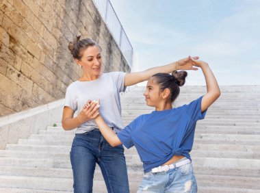 Orta yaşlı anne ve küçük kızı taş merdivenlerde el ele tutuşup birbirlerine bakıyorlar. Mavi tişört ve kot pantolon giyen, yazın dışarıda vakit geçiren bir aile.