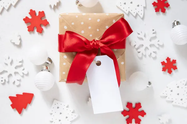 トップビューの周りに白いクリスマスの装飾が施された白いテーブルの上に赤い弓と紙のギフトタグと素朴なラップギフトボックス 空白のギフトタグで冬の組成物 Mockup Copy Space — ストック写真