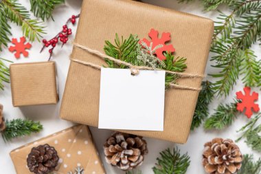 Noel hediyesi olarak, köknar ağacı dalları ve süslemeleri olan beyaz bir masa üzerinde kare kağıt hediye etiketi ile sunulmuştur. Boş hediye etiketli Rustik kış kompozisyonu, boşluğu kopyala