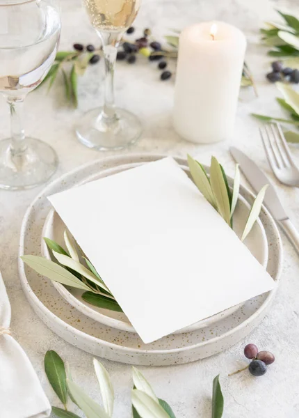 Wedding Table place with a place card and porcelain plates decorated with olive branches close up. Elegant modern template with vertical blank paper card. Mediterranean flat lay mockup, copy space