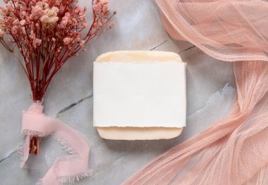 Kağıt ambalaj paketiyle mermer bir masanın üzerinde duran el yapımı sabun, etrafında pembe tül kumaş ve kurumuş çiçekler, üst manzara. Düz sabun, el yapımı yaşam tarzı konsepti.