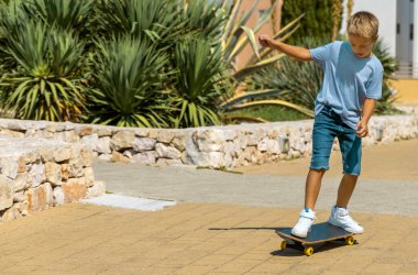 Anaokulu öğrencisi mavi tişörtlü çocuk dışarıda kaykay sürmeyi öğreniyor. Spor yapan ve parkta eğlenen sevimli bir çocuk. Aktif hobiler, sağlıklı yaşam tarzı konsepti. Tişört modeli.