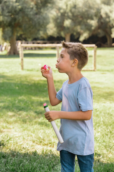 Preschooler boy in blue shirt blowing soap bubbles outdoors. Cute kid boy having fun in a park at summer. Child outdoor activity concept, authentic childhood moment. T-shirt mockup