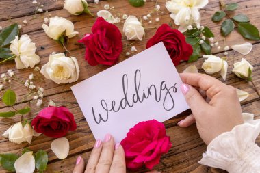 Elle yazılmış evlilik kartı olan eller kırmızı ve kremalı güllerle çevrili, ahşap bir masaya yakın. Çiçeklerin yanında aşk ilanı, Sevgililer Günü konsepti.