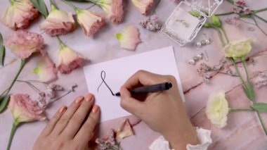 Pembe çiçeklerin yakınındaki bir karta yazı yazan eller mermer bir masaya kapanır.