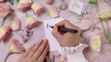 Eller, sen ve beni pembe çiçeklerin yakınındaki bir karta yazıp mermer bir masaya kapatıyor.