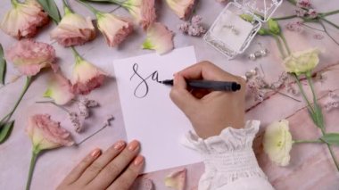 Hands writing SAVE THE DATE on a card near pink flowers close up on a marble table