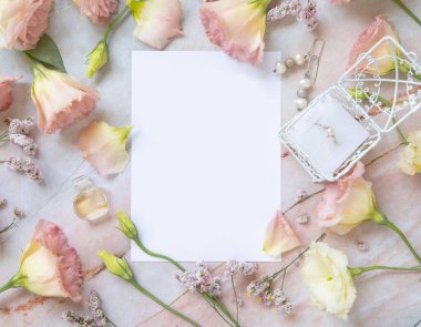 Paper card mockup between pink flowers, petals, earrings and perfume flacon top view on a marble table. Romantic scene with vertical blank card with place for text  flat lay. Valentines, Spring or Mother's day concept
