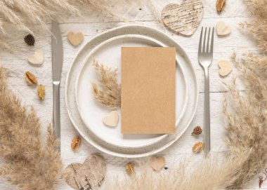 Kurumuş bitkilerin, palmiye yapraklarının, otların ve kalplerin yanında boş kartpostal olan düğün masası. Dikey kraft kağıt kartın düz olduğu boho sahnesi. Selamlama, davetiye ya da sevgililer günü maketi