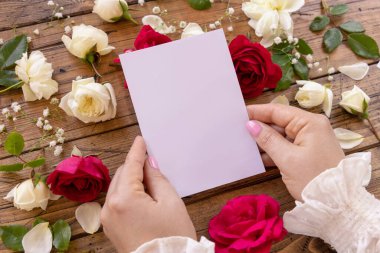 Ellerinde kırmızı ve krema gülleriyle çevrili boş kağıt kart olan eller ahşap bir masanın üzerine kapanır. Çiçeklerin yanında kadın romantizmi, Sevgililer Günü konsepti.