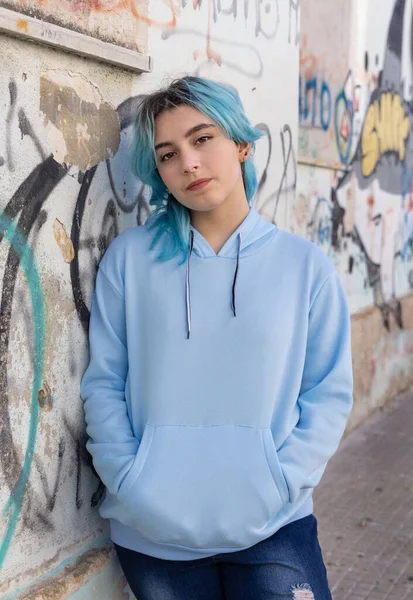 Açık mavi kapüşonlu ve kot pantolonlu bir genç kameraya bakıyor. Mavi saçlı genç kız dışarıda duvar yazısının karşısında duruyor. Giysi modeli. 
