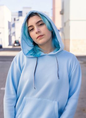 Açık mavi kapüşonlu ve kot pantolonlu bir genç kameraya bakıyor. Mavi saçlı genç kız şehir caddesinde dışarıda kalıyor. Giysi modeli. 