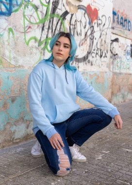 Mavi saçlı, açık mavi kapüşonlu genç kız yan bakıyor. Grafiti duvarının yanında kalçalarının üzerinde oturan mavi saçlı genç kız. Giysi modeli. 