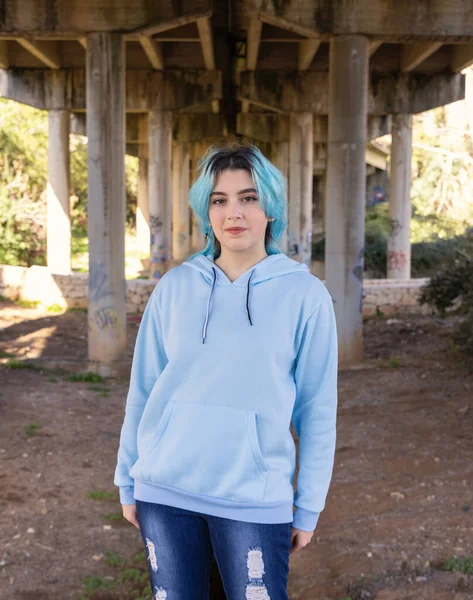Gülümseyen genç açık mavi kapüşonlu ve kot pantolon köprünün altında kalıyor. Mavi saçlı genç kız köprü sütunlarına karşı açık havada kalıyor. Giysi modeli. 