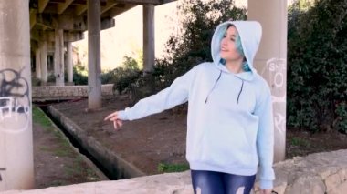 Açık mavi kapüşonlu, kot pantolonlu, gülümseyen ve dans eden bir genç. Mavi saçlı genç kız köprü sütunlarına karşı açık havada kalıyor. Giysi modeli.