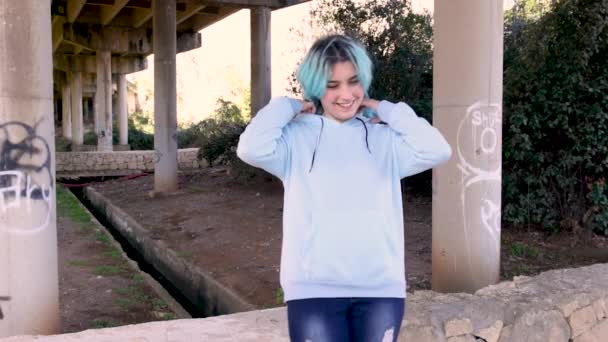 パーカーを着て橋の柱に対して滞在する10代の笑顔 青い髪の10代の女の子は橋の柱に対して屋外にとどまる 服のモックアップ — ストック動画