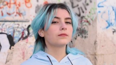 Açık mavi kapüşonlu ciddi bir genç sola döndü. Mavi saçlı genç kız dışarıda grafiti duvarına yaslanıp kameraya bakıyor. Kafasından vur.