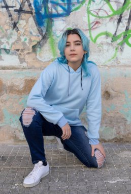 Mavi saçlı, açık mavi kapüşonlu genç kız yan bakıyor. Grafiti duvarının yanında kalçalarının üzerinde oturan mavi saçlı genç kız. Giysi modeli. 