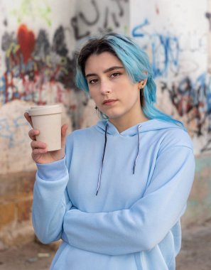 Açık mavi kapüşonlu ve elinde kahve olan genç kız dışarıda grafiti duvarının yanında duran mavi saçlı genç kıza bakmaya gitti. Kıyafet modeli. Hipster ve ergenlik kavramı