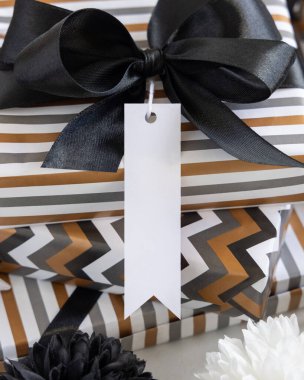 Boş dikey hediye etiketi ve siyah fiyonk var. Çizgili ve simge geometrik kağıda sarılı hediye kutuları, siyah ve beyaz dekorun yanında. Noel, Yeni Yıl, Doğum günü, Yıldönümü Modeli