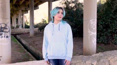 Kapüşonlu giyen ve köprü sütunlarına dayanan gülümseyen genç. Mavi saçlı genç kız köprü sütunlarına karşı açık havada kalıyor. Giysi modeli.
