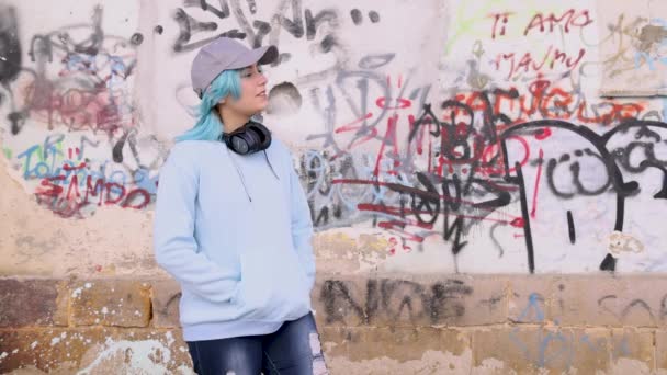 ワイヤレスヘッドフォンとライトブルーのオーバーサイズパーカーのティーンエイジャー 青い髪の10代の女の子は落書きの壁に対して屋外にとどまる 服のモックアップ — ストック動画