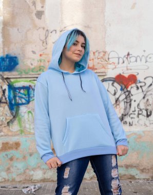 Açık mavi kapüşonlu ve kot pantolonlu bir genç kameraya bakıyor. Mavi saçlı genç kız dışarıda duvar yazısının karşısında duruyor. Kıyafet modeli. Hipster ve ergenlik kavramı