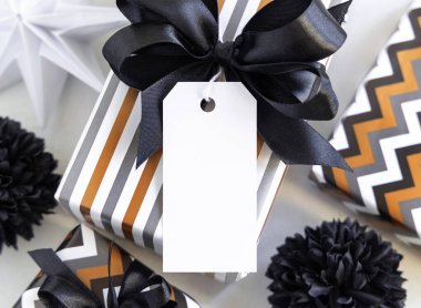 Dikey boş hediye etiketi ve siyah fiyonk var. Siyah beyaz dekorun yanında çizgili geometrik kağıda sarılı hediye kutuları. Noel, Yeni Yıl, Doğum günü, Yıldönümü Modeli