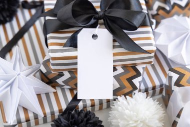 Boş dikey hediye etiketi ve siyah fiyonk var. Çizgili ve simge geometrik kağıda sarılı hediye kutuları, siyah ve beyaz dekorun yanında. Noel, Yeni Yıl, Doğum günü, Yıldönümü Modeli