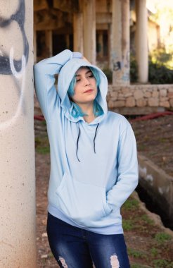 Gülümseyen genç açık mavi kapüşonlu ve kot pantolon köprünün altında kalıyor. Mavi saçlı genç kız köprü sütunlarına karşı açık havada kalıyor. Giysi modeli. 