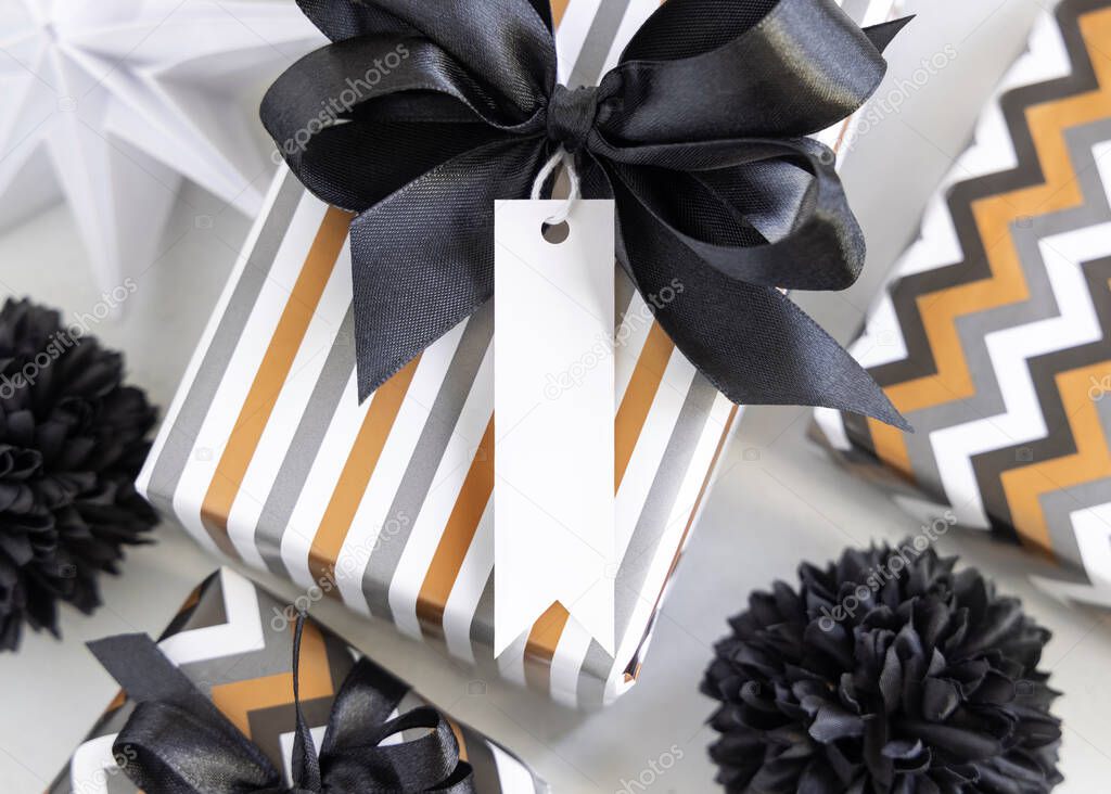 Present with vertical blank gift tag and black bow close up. Gift boxes wrapped in Striped and chevron geometric paper near black and white decor. Christmas, New Year, Birthday, Anniversary label mockup