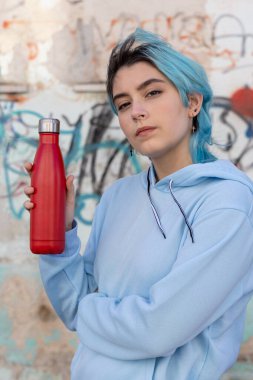 Kırmızı su şişeli mavi kapüşonlu genç. Mavi saçlı genç kız dışarıda grafiti duvarının yanında kalıyor. Giyinme ve tekrar kullanılabilir şişe maketi. Hipster ve ergenlik kavramı