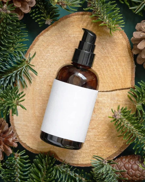 Brown glass bottle between fir branches and pine cones on wooden piece top view. Brand packaging mockup. One pump dispenser for skincare product, cream or serum. Healthcare concept. Natural cosmetics.