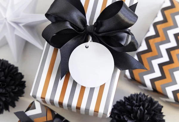 Yuvarlak boş hediye etiketi ve siyah fiyonk var. Siyah beyaz dekorun yanında çizgili geometrik kağıda sarılı hediye kutuları. Noel, Yeni Yıl, Doğum günü, Yıldönümü Modeli