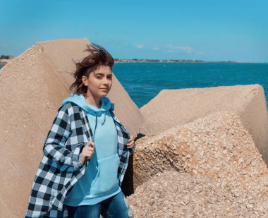 Kısa saçlı, açık mavi kapüşonlu ve ekose gömlekli genç kız kameraya bakıyor. Genç kız dışarıda beton bloklara, deniz ve mavi gökyüzüne, rüzgarlı bir güne karşı durur. Giysi modeli, kopyalama alanı