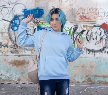 Açık mavi kapüşonlu ve kot pantolonlu gülümseyen genç kameraya bakıyor. Mavi saçlı genç kız dışarıda duvar yazısının karşısında duruyor. Giysi modeli.