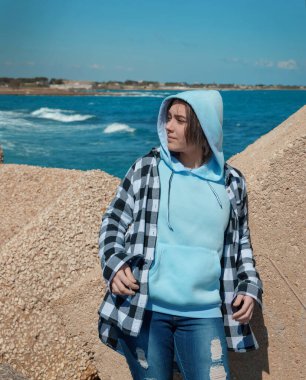 Açık mavi kapüşonlu ve ekose gömlekli genç yan bakıyor. Genç kız deniz kenarında, beton bloklar ve mavi gökyüzü, rüzgarlı bir gün. Giysi modeli, kopyalama alanı