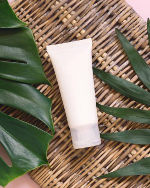 Hasır masa manzarasında, tropik yaprakların arasında duran beyaz plastik boru. Marka ambalaj modeli. Doğal bitkisel kozmetik konsepti - cilt bakımı güzellik ürünü, krem veya orantı