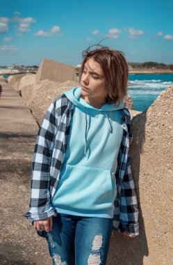 Açık mavi kapüşonlu ve ekose gömlekli genç yan bakıyor. Genç kız deniz kenarında, beton bloklar ve mavi gökyüzü, rüzgarlı bir gün. Giysi modeli, kopyalama alanı