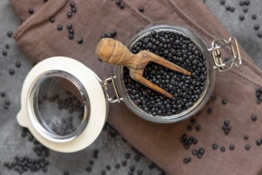 Glass jar of dry black lentils beans with a wooden scoop on grey table top view.   Healthy eating, protein source for vegan and vegetarian diet. Traditional Asian and North African cuisines ingredient