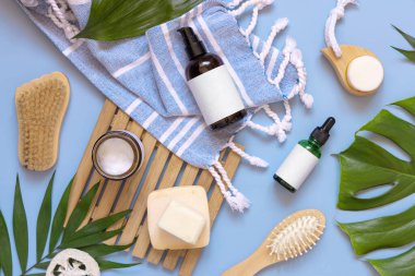 Cam bir pompa makinesi, deri ve saç bakım aksesuarları tropik yaprakların yanında mavi banyo havlusunda, üst manzara, kopyalama alanı. Paketleme modeli. Doğal ev yapımı ürünler