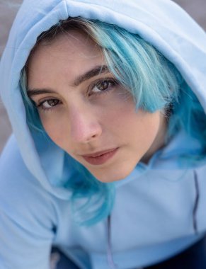 Mavi saçlı, açık mavi kapüşonlu genç kız kameraya yukarıdan bakıyor. Mavi saçlı genç kız açık havada. 