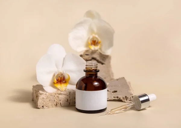 Brown glass dropper bottle on stone near white orchid flowers on light yellow, close up, mockup. Skincare handmade beauty product, serum or lotion. Exotic natural cosmetic