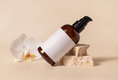 Brown glass one pump bottle near white orchid flower and beige stones on light yellow close up, mockup. Skincare beauty product package. Natural cosmetics, minimal compositio