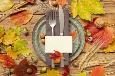 Autumn rustic table place with blank card between colorful leaves and berries, top view, mockup. Flat lay with horizontal card on wooden table,  copy space. Thanksgiving or harvest concept