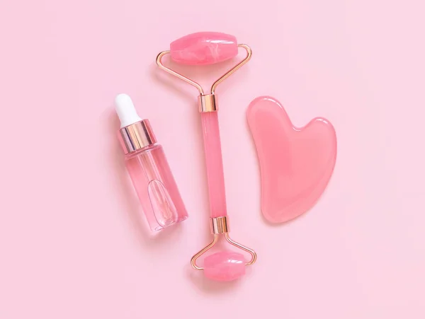 Facial roller, gua sha massager and serum bottle near white orchid flowers on pink top view. Anti-age massage and natural lifting therapy at home. Facial skin care at home. Beauty concept