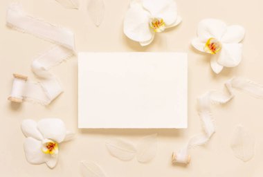 Wedding Card near white orchid flowers and silk ribbons on light yellow top view, mockup. Romantic scene with horizontal blank card flat lay. Valentines or Spring romantic concept